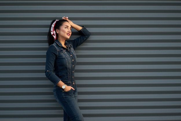 Retrato bastante morena chica de moda joven con el pelo largo, sonrisa encantadora y labios pintados de rojo vistiendo overoles de mezclilla moderna hipster posando sobre fondo gris de la pared urbana.