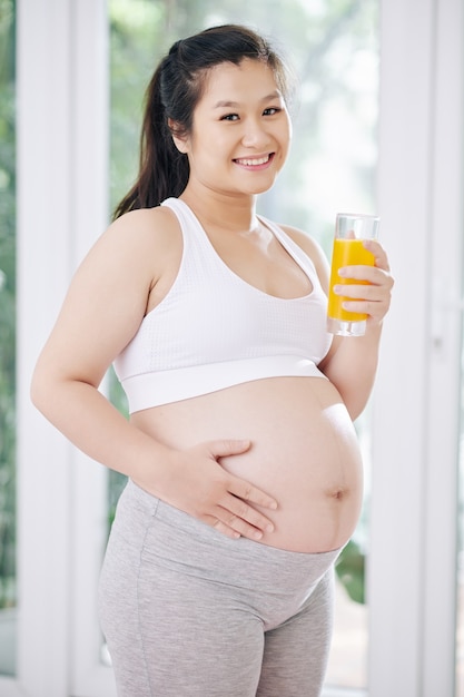 Retrato de bastante sonriente joven mujer embarazada vietnamita bebiendo un vaso de jugo de naranja y tocando el vientre