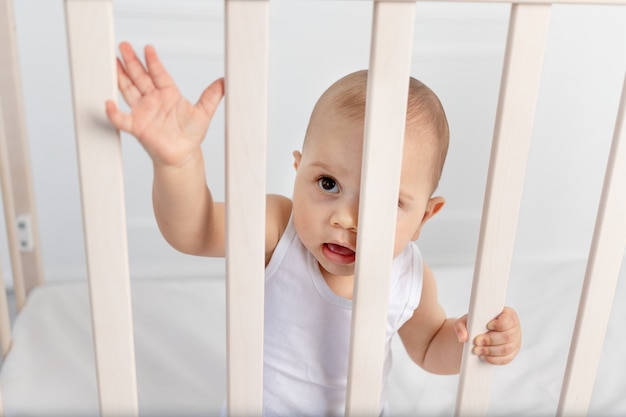 Retrato de un bebé de 8 meses de pie en una cuna en una habitación de niños con ropa blanca y mirando a través de la cama, la mañana del bebé, concepto de productos para bebés