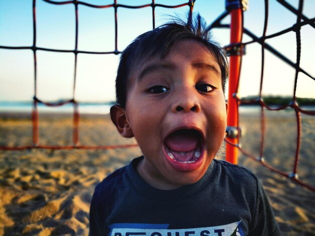 Foto retrato de un bebé con la boca abierta de pie en la playa contra el cielo durante la puesta de sol