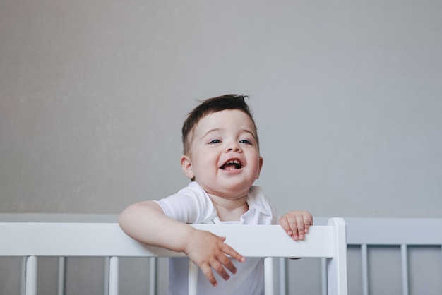 Retrato de un bebé niño gateando y sonriendo en traje blanco en la cama en el dormitorio. Ojos grandes azules Concepto de infancia feliz con vista lateral copyspace