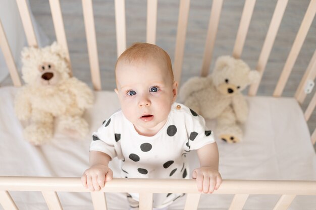 Retrato de un bebé de pie en una cuna con juguetes en pijama después de dormir