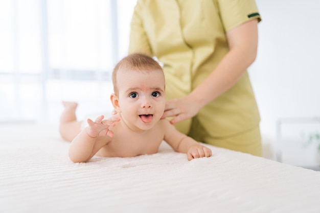 Retrato de un bebé recién nacido sonriente acostado sobre el vientre mientras una masajista femenina irreconocible realiza un masaje profesional para la espalda desarrollando músculos Las manos de la madre hacen un masaje en la espalda al hijo recién nacido
