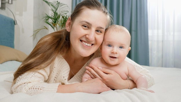 Foto retrato de bebé sonriente y madre acostada en la cama y mirando en cámara