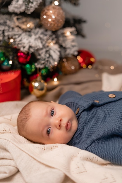 El retrato de un bebé con un traje azul de punto está sobre una manta ligera en el suelo debajo del árbol de Navidad. primera navidad. vacaciones de invierno, momentos divertidos, navidad infantil. foto de alta calidad