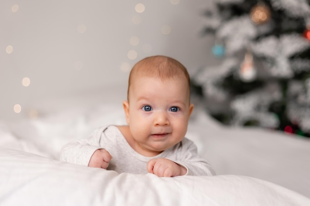 Retrato de un bebé con un traje blanco acostado boca abajo contra el fondo del árbol de Navidad