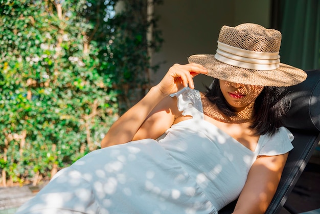 Retrato bela viajante asiática de vestido branco e chapéu de palha relaxando na cadeira de praia à beira da piscina Conforto resort férias de verão