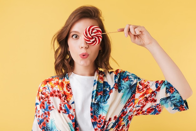 Retrato de una bella dama con una camisa colorida parada y cubriendo su ojo con caramelos de piruleta mientras mira con asombro a la cámara sobre un fondo rosa