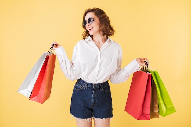 Retrato de una bella dama sonriente con gafas de sol de pie con coloridas bolsas de compras en las manos sobre fondo rosa Mujer bonita de pie con camisa blanca y pantalones cortos de mezclilla