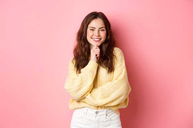 Retrato de una bella dama sonriente, luciendo determinada, tocando la barbilla y mirando satisfecha, viendo algo interesante, tengo una idea, de pie pensativo contra la pared rosa.