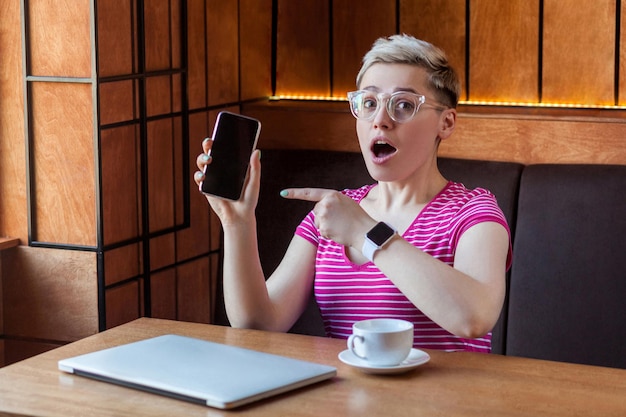 El retrato de una bella e increíble joven bloguera con el pelo corto en pantalones rosa y anteojos está sentado en un café, sosteniendo el teléfono y señalando con el dedo, mirando a la cámara con la cara sorprendida. Interior,