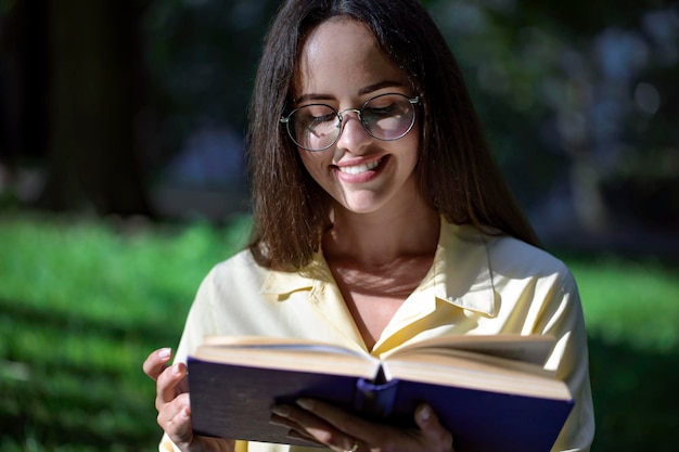 Retrato de una bella estudiante morena con gafas leyendo un libro al aire libre