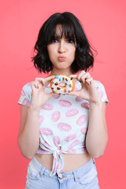 Retrato de una bella joven caucásica vestida con una camiseta con donas mostrando una rosquilla y mostrando una cara gorda sobre un fondo rosa.