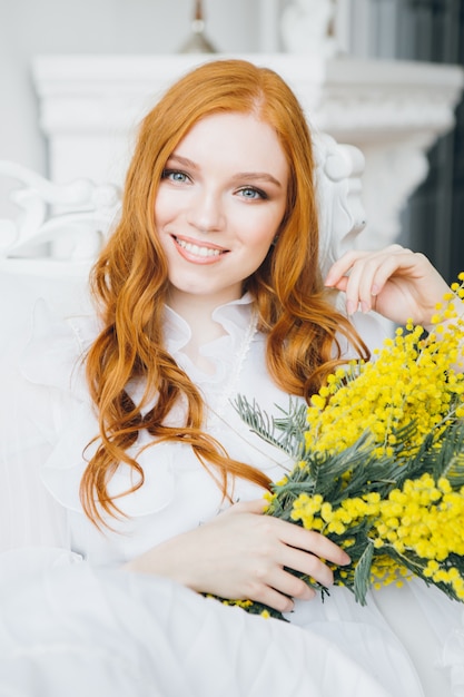 Retrato de una bella joven pelirroja con una mimosa en un vestido blanco largo