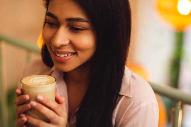 Retrato de una bella joven relajada sosteniendo un vaso de sabroso café con leche en sus manos y sonriendo