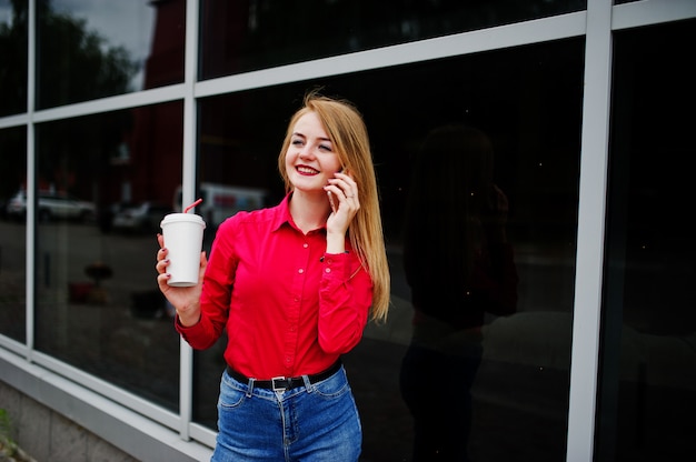 Retrato de una bella mujer en blusa roja y jeans casuales hablando por teléfono móvil y sosteniendo una taza de café fuera del enorme centro comercial.