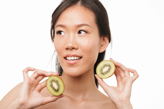 Retrato de una bella mujer bonita joven asiática positiva con piel sana posando aislada sobre pared blanca sosteniendo kiwi