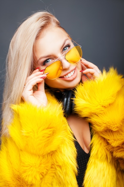 Retrato de una bella mujer joven con auriculares en gafas de sol de moda amarillo con un abrigo de moda amarillo sobre gris