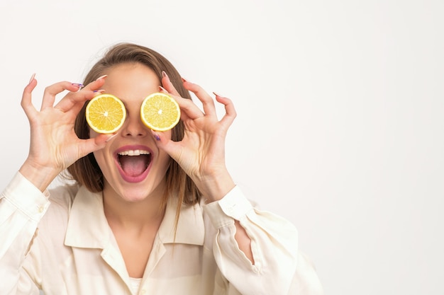 Retrato de una bella mujer joven con la boca abierta sosteniendo dos rodajas de naranja en sus ojos sobre un fondo blanco.