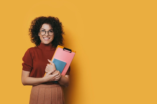 Retrato de una bella mujer joven con cabello rizado con gafas mirando a la cámara riendo mientras sostiene sus libros de estudio aislados en la pared amarilla del estudio.