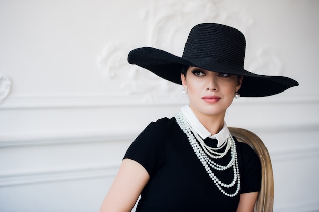 Retrato de una bella mujer joven en estilo retro con un elegante sombrero negro y vestido sobre pared rococó de lujo