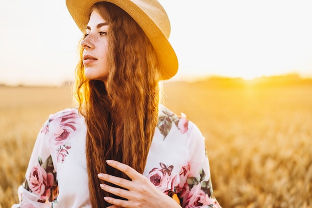 Retrato de una bella mujer joven con pelo rizado y cara de pecas. Mujer en vestido y sombrero posando en el campo de trigo al atardecer y mirando