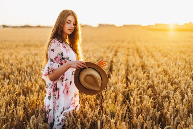 Retrato de una bella mujer joven con pelo rizado y cara de pecas. Mujer en vestido y sombrero posando en el campo de trigo al atardecer y mirando