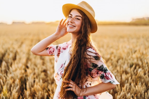 Retrato de una bella mujer joven con pelo rizado y cara de pecas. Mujer en vestido y sombrero posando en el campo de trigo al atardecer y mirando