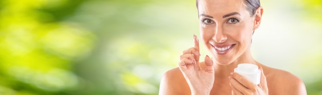 Retrato de una bella mujer joven con piel muy suave sosteniendo una crema blanca. Concepto de cosmética y bienestar de cuidado de la piel de spa. Bandera panorámica verde borrosa.