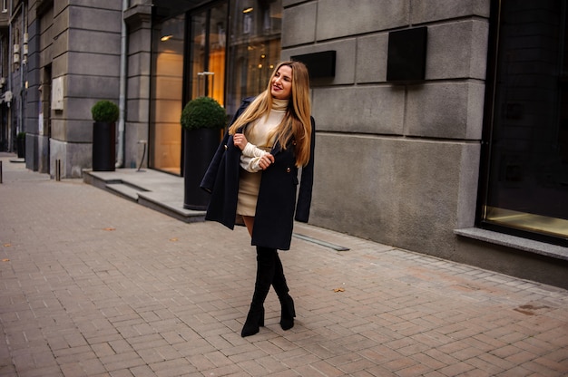 Retrato de una bella mujer de pie en la acera con vestido beige y abrigo negro