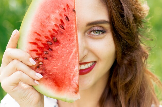 Retrato de una bella mujer sonriente sosteniendo una rodaja de sandía que cubre la mitad de la cara