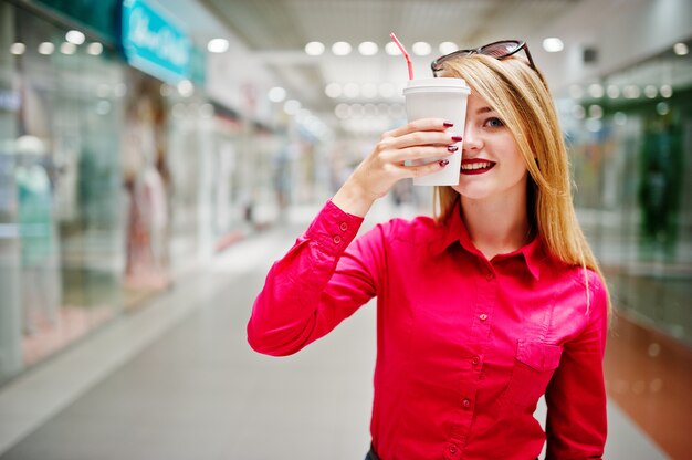 El retrato de una bella mujer vestida con una blusa roja, jeans casuales y tacones negros cubre la mitad de su rostro con una taza de café en un enorme centro comercial.