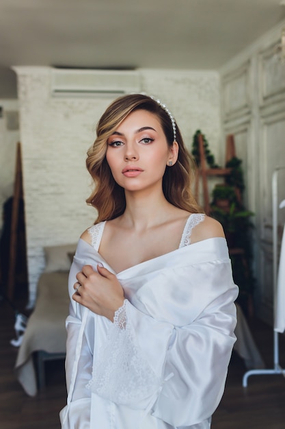 Retrato de una bella novia rubia en el interior. Novia joven de la mañana en una bata de baño. Fotografía de boda. Niña sonriente y linda se encuentra.