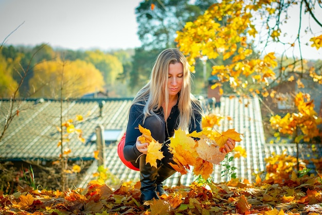 Retrato de una bella rubia con una falda corta roja recogiendo follaje amarillo en el parque de otoño.