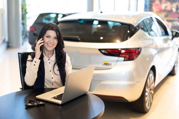 Retrato de bella smilling agente de seguros sentado y hablando por teléfono cerca de un auto nuevo