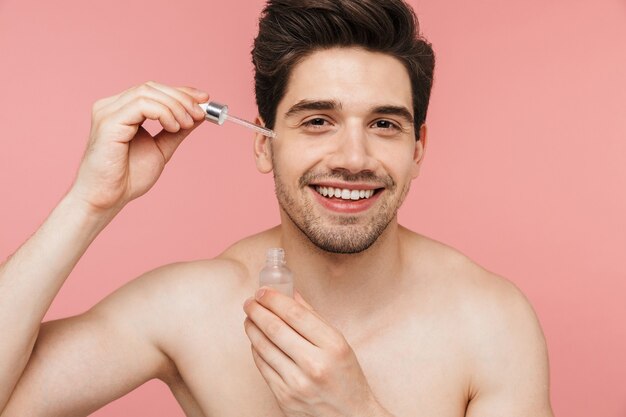 Retrato de belleza de un apuesto hombre morena sin camisa sonriente que se encuentran aisladas sobre la pared rosa, aplicando aceite facial de una botella