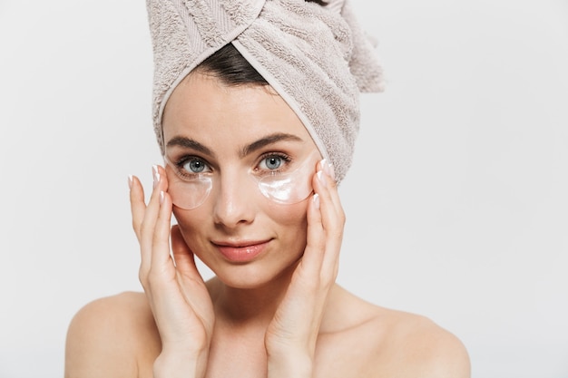 Retrato de belleza de una atractiva joven morena de pie aislado sobre una pared blanca, con una toalla en la cabeza, aplicando parches cosméticos en los ojos