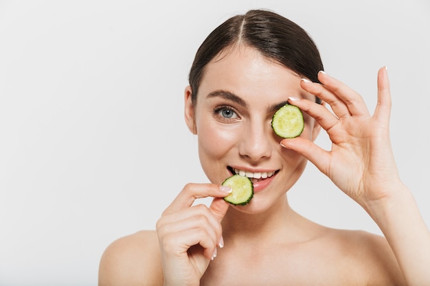 Retrato de belleza de una atractiva joven morena que se encuentran aisladas sobre una pared blanca, sosteniendo pepino en rodajas