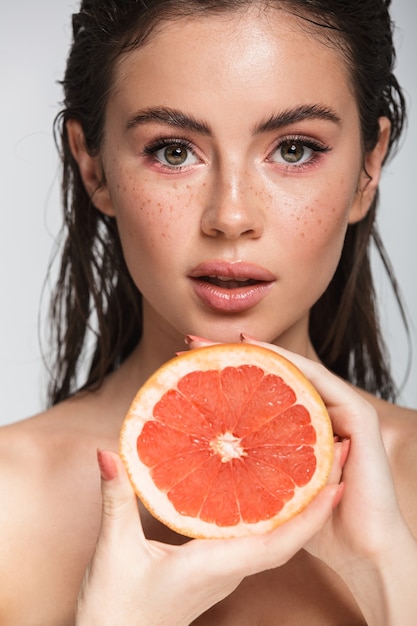 Foto retrato de belleza de una atractiva joven mujer morena en topless de pie aislado en blanco, posando con pomelo partido a la mitad