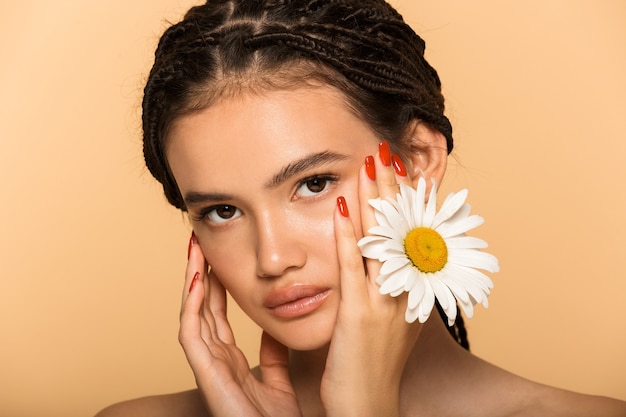 Retrato de belleza de una atractiva joven en topless que se encuentran aisladas sobre la pared beige, posando con flor de manzanilla