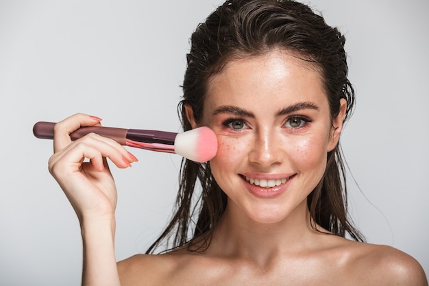 Retrato de belleza de una atractiva mujer joven sensual sonriente con pelo largo morena mojado de pie aislado en gris, con pincel de maquillaje