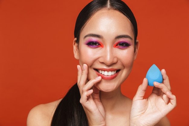 Retrato de belleza de una feliz joven mujer asiática en topless con cabello morena con maquillaje brillante, que se encuentran aisladas en rojo, sosteniendo una esponja de maquillaje