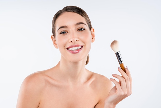 Retrato de belleza de una joven mujer sonriente sosteniendo un cepillo de maquillaje aislado sobre fondo blanco