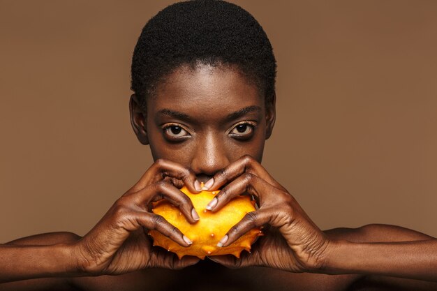 Retrato de belleza de mujer africana semidesnuda bastante joven con melón de cuernos kiwano aislado en marrón