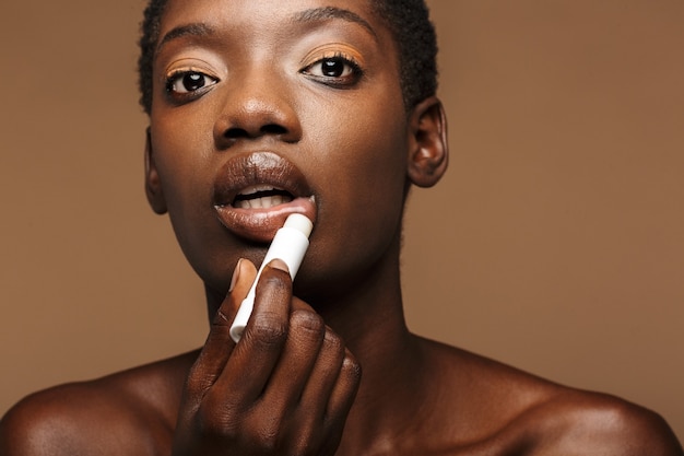 Retrato de belleza de mujer africana semidesnuda bastante joven poniéndose bálsamo labial aislado en marrón