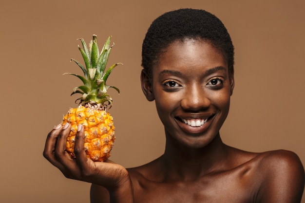 Retrato de la belleza de la mujer africana semidesnuda bastante joven que sostiene la piña aislada en marrón