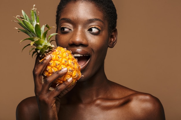 Foto retrato de la belleza de la mujer africana semidesnuda bastante joven que sostiene la piña aislada en marrón