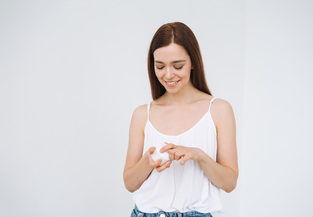 Retrato de belleza de una mujer feliz y sonriente con cabello largo y oscuro que pone crema humectante nutritiva para el día en la cara y las manos limpias y frescas de la piel en el fondo blanco aislado