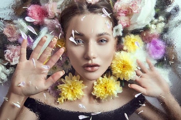 Retrato de belleza de una mujer con flores y pétalos detrás del cristal en gotas de lluvia