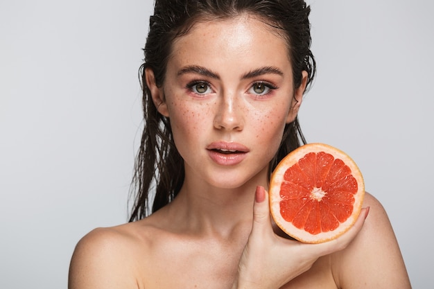 Retrato de belleza de una mujer joven sensual atractiva con el pelo largo morena mojado de pie aislado en gris, mostrando pomelo partido a la mitad, posando